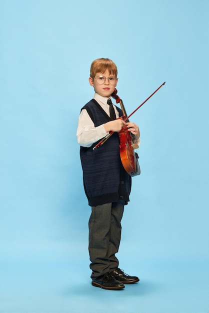 Niño lindo y serio pequeño músico con ropa de gran tamaño posando con violín aislado sobre azul