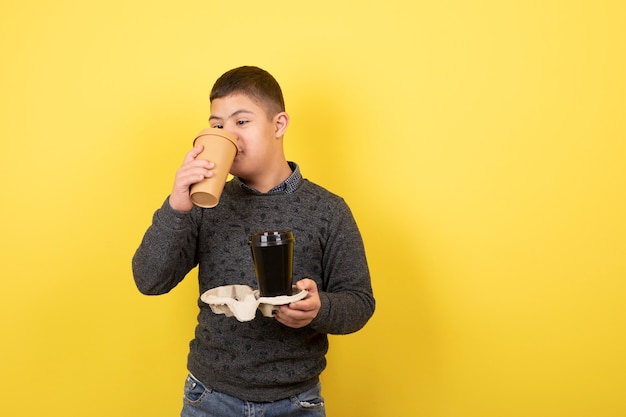 niño lindo con síndrome de down bebiendo una taza de café sobre amarillo.