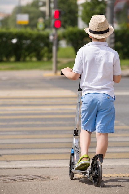 Niño lindo con sombrero de paja en un scooter parado en una encrucijada y esperando que la luz se ponga verde Niño esperando en luz verde en la carretera Colegial en scooter cruzando la calle en un día soleado