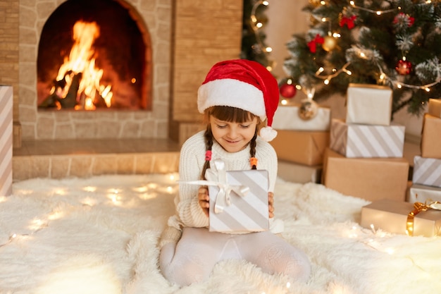 Niño lindo con sombrero rojo festivo con caja de regalo de Navidad posando en casa mientras está sentado en el piso sobre una alfombra suave cerca de la chimenea y el árbol de Navidad. Niña encantadora con coletas tiene caja de regalo.