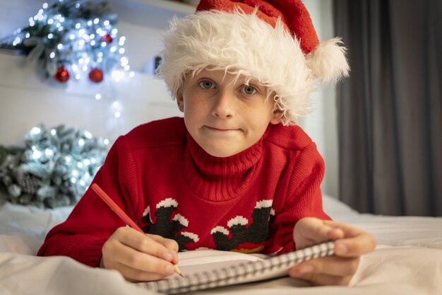 Niño lindo con sombrero de Santa Claus escribe una carta a Santa Claus para obtener un regalo acostado en la cama con el telón de fondo de las decoraciones navideñas en casa. Navidad, año nuevo 2022, Santa Claus. Deseo. milagro.