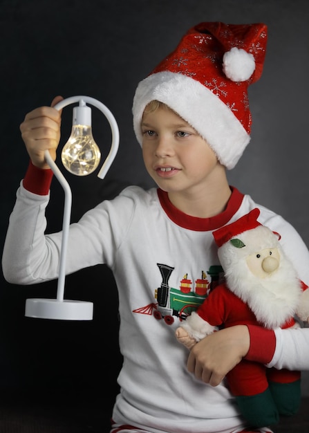 Niño lindo con sombrero de santa sosteniendo la linterna y un pequeño santa sobre fondo oscuro, Navidad