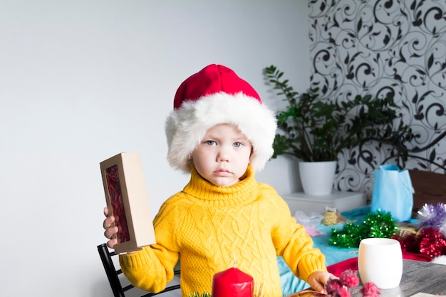 Un niño lindo con un suéter amarillo y un sombrero rojo de Papá Noel en una mesa de madera con regalos empacados en papel kraft