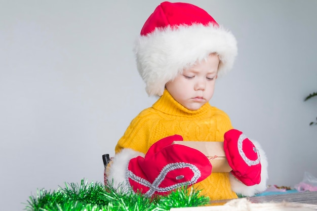 Un niño lindo con un suéter amarillo, un sombrero rojo de Papá Noel y mitones de Navidad con regalos en las manos