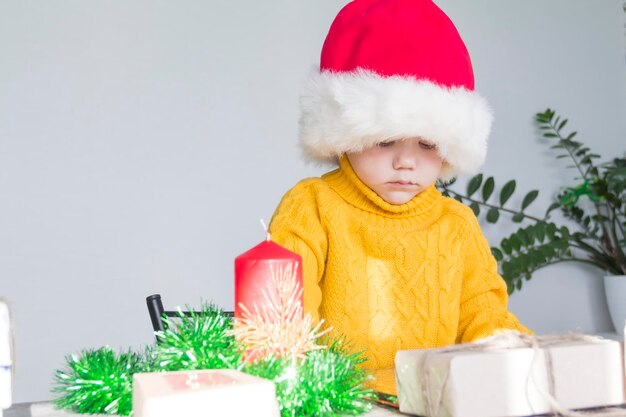 Foto un niño lindo en un suéter amarillo y un sombrero rojo de santa claus en una mesa de madera con regalos que