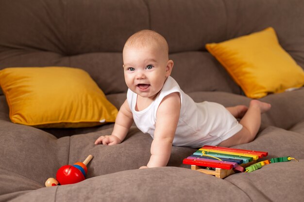 Niño lindo en traje blanco se encuentra en casa en un sofá gris con almohadas amarillas jugando con juguete de madera en desarrollo