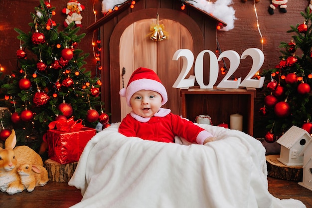 Niño lindo vestido como Santa Claus se sienta cerca del árbol de Navidad. productos para niños y vacaciones. Concepto de año nuevo de invierno. navidad 2022