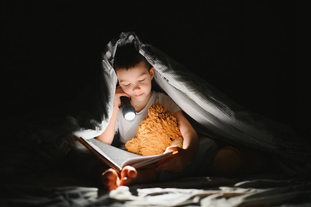 Niño con linterna leyendo un libro debajo de una manta en casa