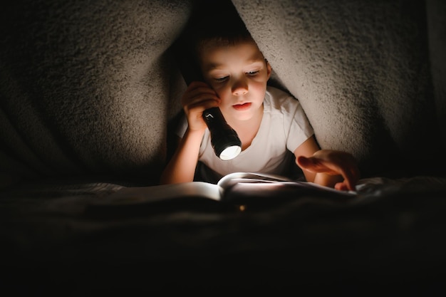 Niño con linterna leyendo un libro debajo de una manta en casa