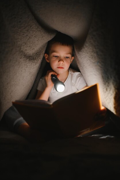 Niño con linterna leyendo un libro debajo de una manta en casa