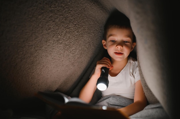 Niño con linterna leyendo un libro debajo de una manta en casa