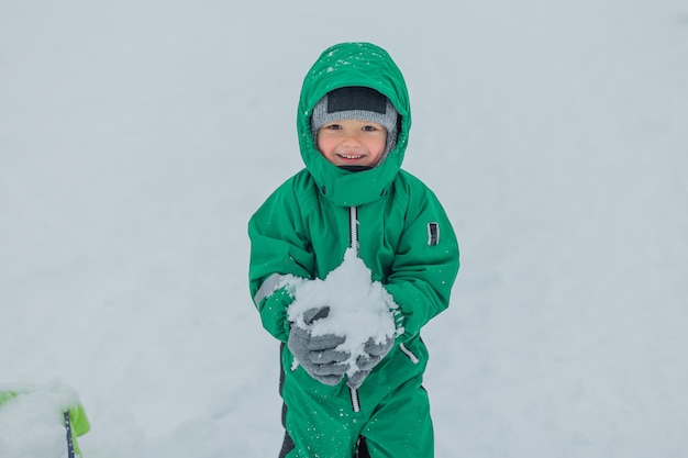 Foto el niño lleva un trozo de nieve en sus manos y sonríe. el niño está vestido con un mono verde cálido.