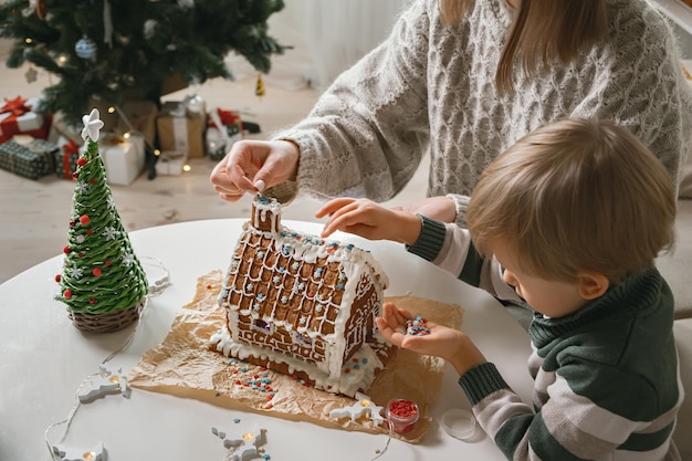 Niño con mamá decorando juntos la casa de pan de jengibre de Navidad, actividades familiares y tradiciones en Navidad y Nochevieja