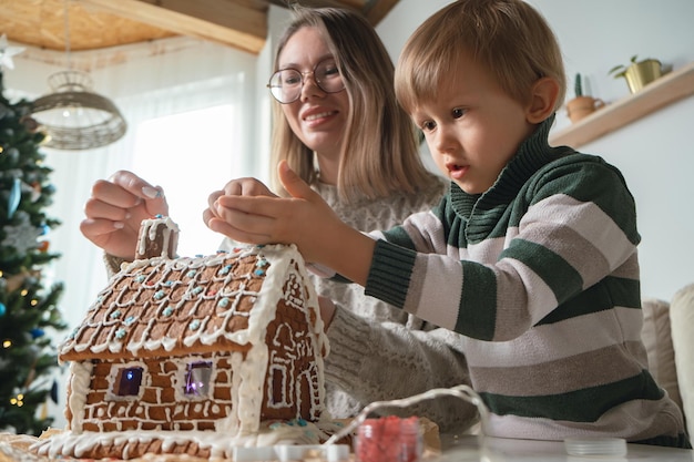 Niño con mamá decorando juntos la casa de pan de jengibre de Navidad, actividades familiares y tradiciones en Navidad y Nochevieja