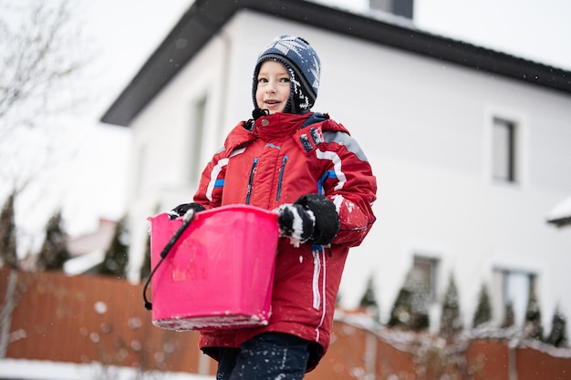 Niño mantenga cubo con nieve contra casa en invierno activo al aire libre ocio infantil