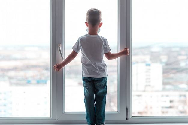 Un niño con una máscara médica está sentado en su casa en cuarentena debido a coronavirus y covid -19 y mira por la ventana.