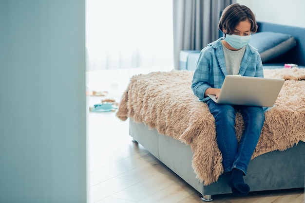 Niño con máscara médica sentado en la cama con una computadora portátil en su regazo Banner de plantilla