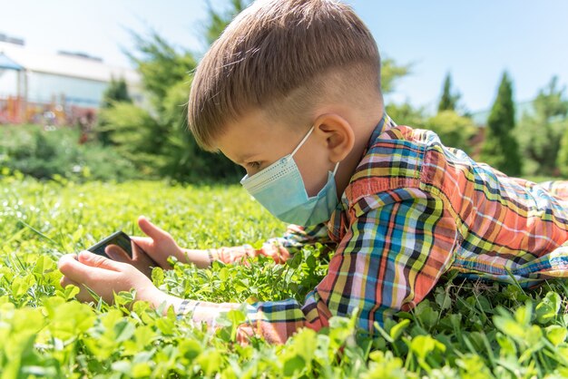 Un niño con una máscara médica se sienta en la hierba y mira en el teléfono
