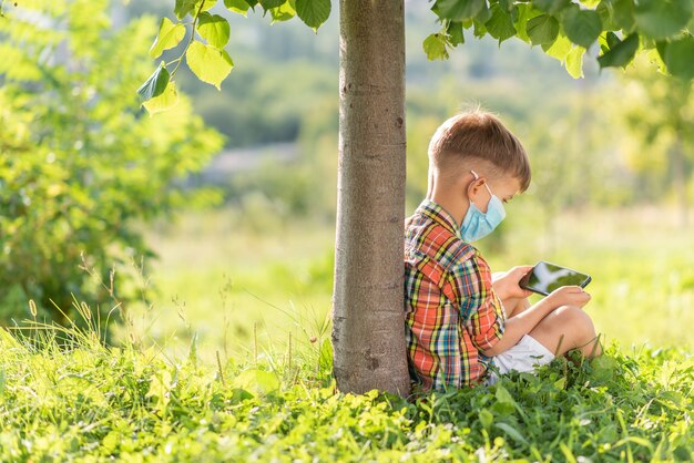 Un niño con una máscara médica se sienta en la hierba y mira en el teléfono