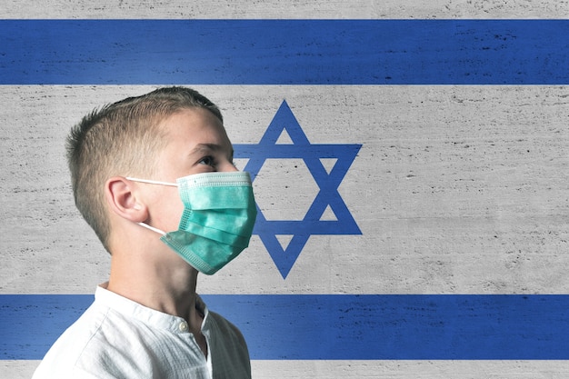 Niño con una máscara médica en su rostro en el fondo de la bandera de Izrael.