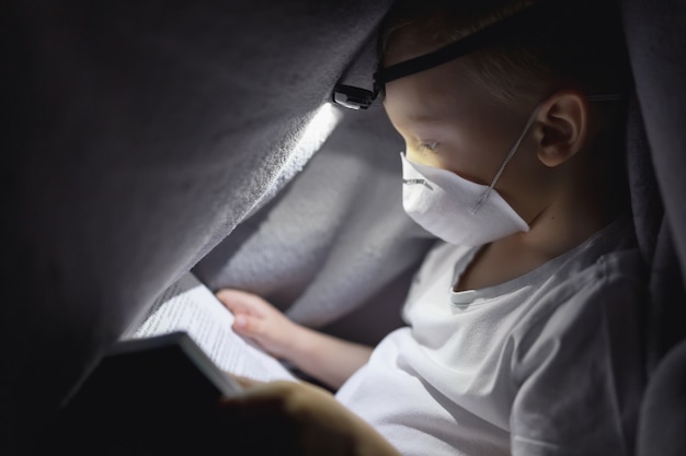 Un niño con una máscara protectora debajo de una manta leyendo un libro con una linterna. El concepto de pasar tiempo en aislamiento seguro.