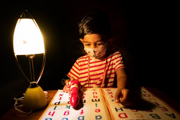 Un niño con una máscara de seguridad está sentado en casa leyendo un libro de aprendizaje del alfabeto inglés Niño asiático estudiando bajo la lámpara Foto de concepto educativo infantil