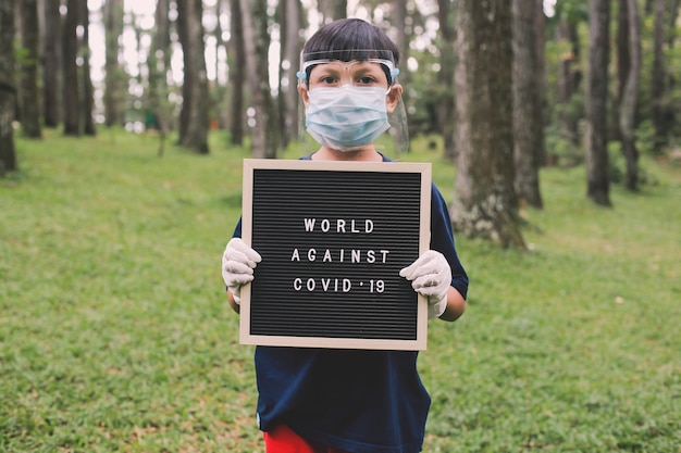 Niño con mascarillas y guantes de goma de pie y sosteniendo el tablero de letras que escrito World Against Covid