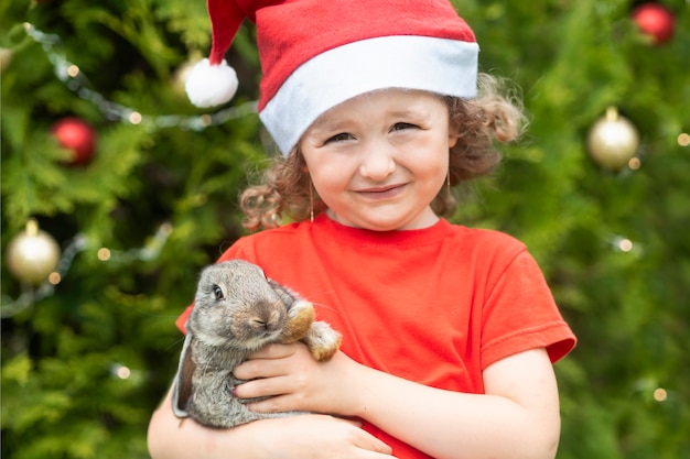 Niño y mascota bajo el árbol de Navidad. niña feliz con gorro de Papá Noel con conejito de regalo, conejo para año nuevo