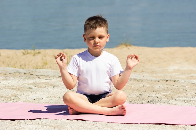 Niño medita sosteniendo los dedos en pose de yoga con los ojos cerrados sentado en una estera en la playa