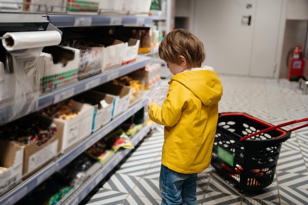 Niño en el mercado con un carrito de supermercado pone dulces en una bolsa