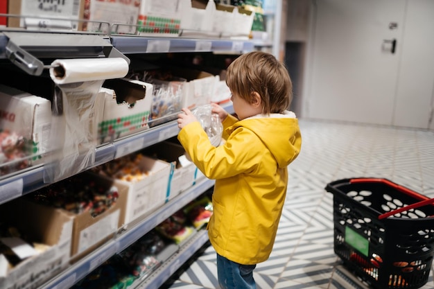 Niño en el mercado con un carrito de supermercado pone dulces en una bolsa