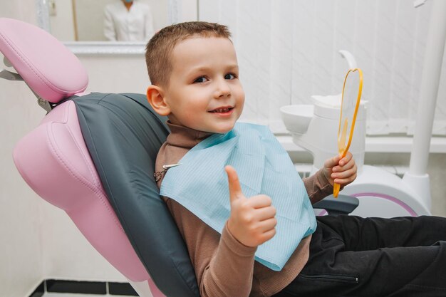 Niño se mira en el espejo con una gran sonrisa, sentado en una silla con un dentista en el consultorio dental.