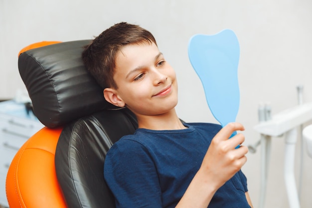 El niño se mira en el espejo con una sonrisa con dientes sentado en una silla con un dentista en el consultorio dental