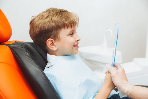 El niño se mira en el espejo con una sonrisa con dientes sentado en una silla con un dentista en el consultorio dental