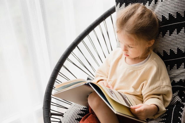 El niño mira las páginas de un libro sentado en una silla cómoda en el balcón del apartamento durante el día