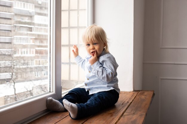 El niño mira por la ventana. Adorable niño rubio sentado en el alféizar de la ventana.