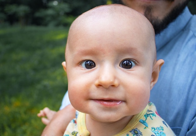 Niño mirando a la cámara y sonriendo de cerca. Papá tiene a su hijo en brazos al aire libre.