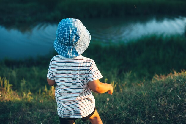 Niño mirando un pequeño río y arrojando piedras con un sombrero azul en un campo verde