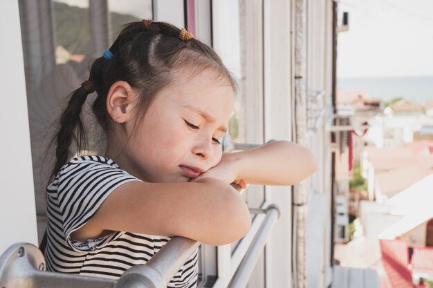 Un niño molesto se para junto a una ventana abierta, el niño se enfermó durante las vacaciones y está triste una niña de asiático ap ...