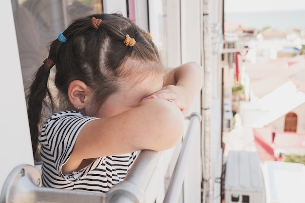 Un niño molesto se para junto a una ventana abierta, el niño se enfermó durante las vacaciones y está triste una niña de asiático ap ...