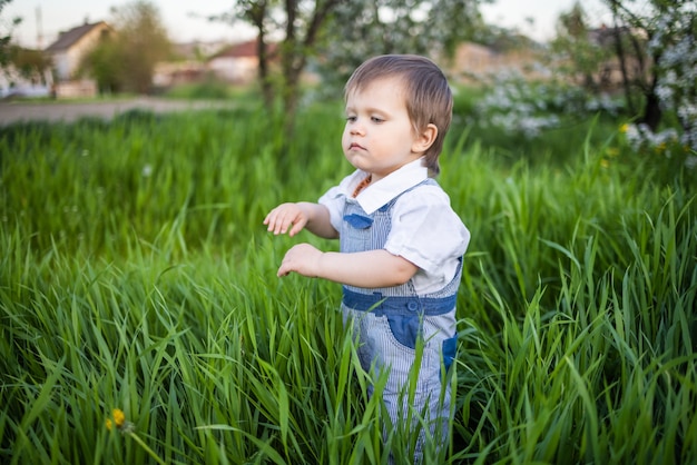 Un niño con un mono de mezclilla con expresivos ojos azules. Saltando y jugando en la hierba verde alta con el telón de fondo de un gran arbusto verde y un jardín floreciente.