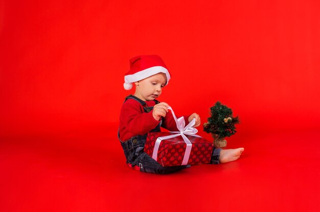 Niño con un mono de mezclilla y un sombrero de Navidad con un pequeño árbol de Navidad se sienta