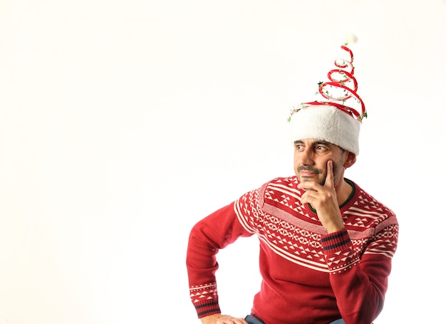 Niño morena sentado en suéter rojo y sombrero de navidad sobre fondo blanco.