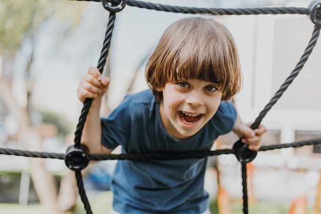 Un niño moreno bonito con una camiseta azul está escalando una red de cuerdas. el niño se dedica a los deportes al aire libre. niño sano y activo. estilo de vida. espacio para texto. foto de alta calidad