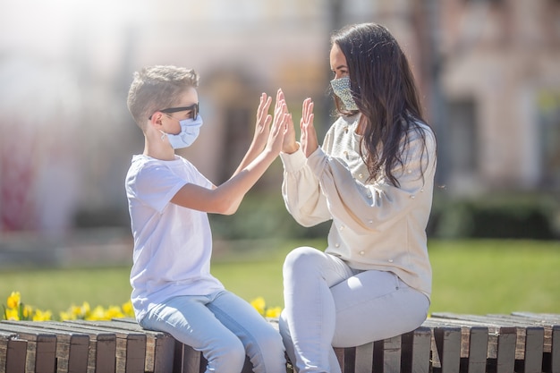 Niño y mujer joven dando un alto cinco con ambas manos sentadas en el exterior, máscaras faciales weraing.