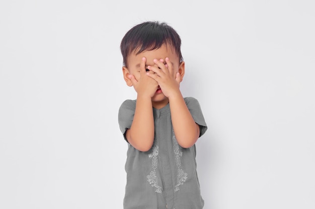 Un niño musulmán asiático sonriente y atractivo que cubre su rostro con la mano aislado en el fondo del estudio blanco