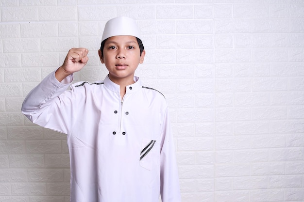 niño musulmán vestido con túnica blanca y gorra levantando los brazos derechos y señalando con el dedo índice hacia arriba