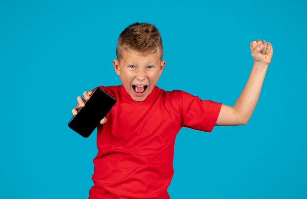 Niño muy contento sosteniendo un teléfono inteligente con pantalla en blanco y celebrando el éxito