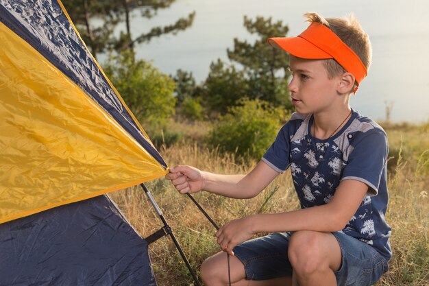 Niño en la naturaleza, en el bosque monta una tienda de campaña y equipa un campamento, en el fondo un lago o un mar, un viaje de senderismo de verano, un campamento