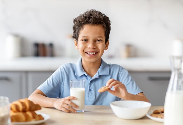 Foto un niño negro feliz bebiendo leche y comiendo galletas en la cocina.